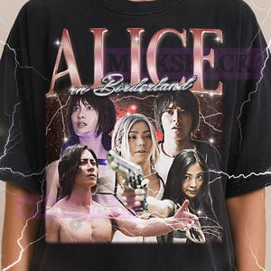 以下が含まれることがあります： 「アリス・イン・ボーダーランド」のグラフィックがプリントされた黒いTシャツ。タイトルはかすれたフォントで、登場人物の画像が描かれています。上部にはMilkshakeのロゴ。ヴィンテージスタイルのデザイン。