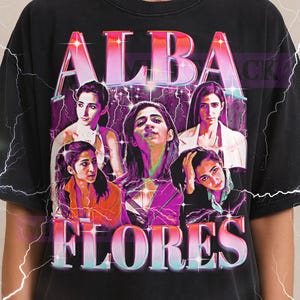 May include: Black t-shirt featuring the text "ALBA FLORES" in a pink and blue gradient, with a graphic of several women. The Milkshake logo is at the top. The shirt is a unisex size.
