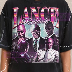 May include: Black t-shirt featuring a graphic of Lance Reddick with his name in a pink and purple design. The shirt has a vintage look and a crew neck.