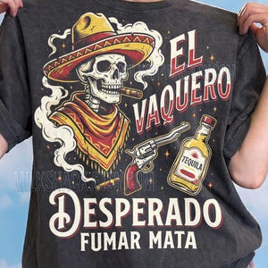 Könnte beinhalten: Dunkelgraues T-Shirt mit einem Skelett-Cowboy-Motiv, das einen Sombrero trägt, eine Zigarre raucht und eine Pistole hält. Das Shirt zeigt den Text "El Vaquero" und "Desperado Fumar Mata" sowie eine Tequila-Flasche.