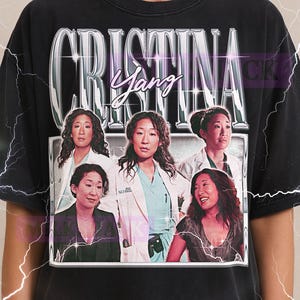 May include: Black t-shirt featuring a graphic of the character Cristina Yang from the TV show Grey's Anatomy. The design includes the name "Cristina Yang" in a silver, stylized font, and multiple images of the character. The Milkshake logo is at the top.