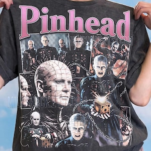 May include: A charcoal gray t-shirt featuring a large pink 'Pinhead' graphic at the top. The shirt displays a collage of multiple scenes of the character Pinhead from the Hellraiser film series, including images of the character holding a puzzle box.