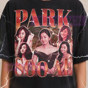 Puede incluir: Camiseta negra con un diseño gráfico retro con el texto "PARK SOO-AH" e imágenes de una mujer. El logotipo de Milkshake está en la parte superior. La camiseta tiene un aspecto vintage.