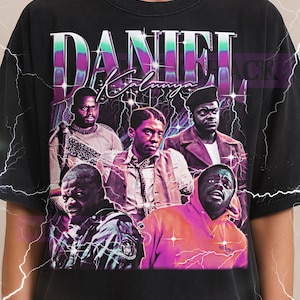 Puede incluir: Camiseta unisex negra con un gráfico de Daniel Kaluuya y otros actores. El diseño incluye el nombre "DANIEL KALUUYA" en una fuente estilizada con acentos de rayos. El logotipo de Milkshake está en la parte superior.