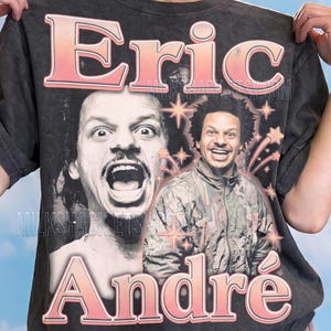 May include: A charcoal gray short-sleeve t-shirt featuring a graphic print of Eric André. The design includes the name Eric André in large pink gradient lettering, two black and white portraits of the comedian, and pink starburst accents.