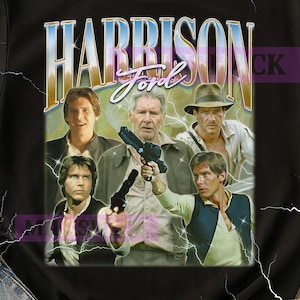 May include: Black t-shirt featuring a graphic of Harrison Ford in various roles, including Han Solo and Indiana Jones. The design includes the name "Harrison Ford" in a retro font with a light blue and gold color scheme. The shirt has a vintage aesthetic.