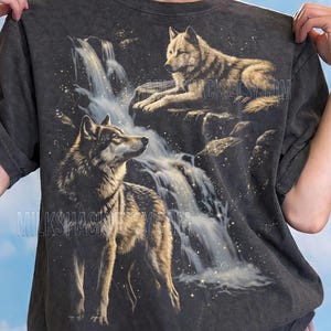 May include: Dark grey t-shirt featuring a detailed illustration of two wolves near a waterfall. The design includes the text "MILKSHACKETSY.COM" and "Comfort Colors: Pepper". The shirt is designed by MILKSHACK.