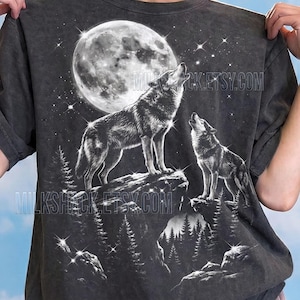 May include: Dark grey t-shirt featuring a detailed graphic of two wolves howling at a full moon in a starry night sky. The design includes pine trees and rocky terrain. The text "Comfort Colors: Pepper" is at the bottom.