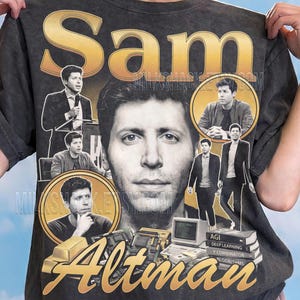 Op de afbeelding: Een antracietgrijs t-shirt met een grafische print van Sam Altman. Het ontwerp bevat meerdere fotografische portretten van Altman, de tekst "Sam" en "Altman" in goud, en illustraties van computerhardware en een stapel boeken.
