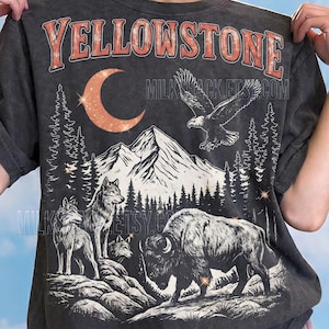 Puede incluir: Camiseta gris oscuro con un diseño gráfico desgastado. El diseño incluye la palabra "YELLOWSTONE" en naranja, una luna creciente, un águila, montañas, árboles, lobos y un bisonte. El texto "Comfort Colors: Pepper" está en la parte inferior.