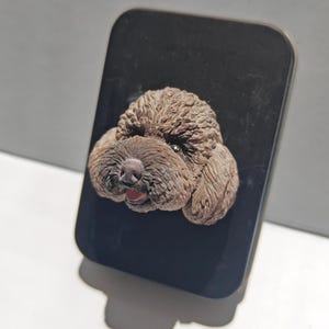 May include: A brown poodle head sculpture is attached to a black rectangular surface. The poodle's fur is textured, and its mouth is slightly open, revealing a pink tongue. The sculpture is set against a white background.