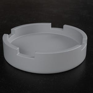May include: A light gray ashtray with a circular design. The ashtray has a low profile with a recessed center and four notched areas around the rim. The ashtray is made of a solid, smooth material and sits on a dark surface.