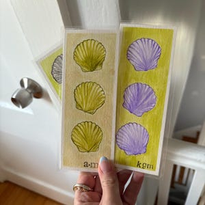 May include: Three watercolor bookmarks featuring seashell designs. Two bookmarks have three seashells each, one in yellow and the other in purple. The third bookmark has a single gray seashell. The initials "a.m" and "kgm" are printed on the bookmarks.