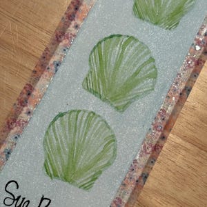 May include: A rectangular bookmark with a light blue glittery surface featuring three green seashell illustrations arranged vertically. The edges are decorated with a floral patterned border, and the text Sue Beach is written in black script.