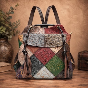Può includere: Uno zaino patchwork multicolore con motivi floreali in rilievo nei toni del rosso, verde, marrone e nero. La borsa presenta una patta superiore, due spallacci marrone scuro e due lunghe nappe in pelle marrone pendenti sul davanti.