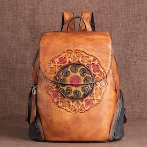 May include: A brown leather backpack with a decorative floral design on the front flap. The backpack features a top handle, adjustable straps, and side zips. The floral design incorporates red, green, and yellow accents.