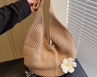 May include: A beige, open-weave tote bag with a single handle. A white crocheted flower with a yellow center is attached to the side of the bag. The bag is held by a person wearing a black sleeve.