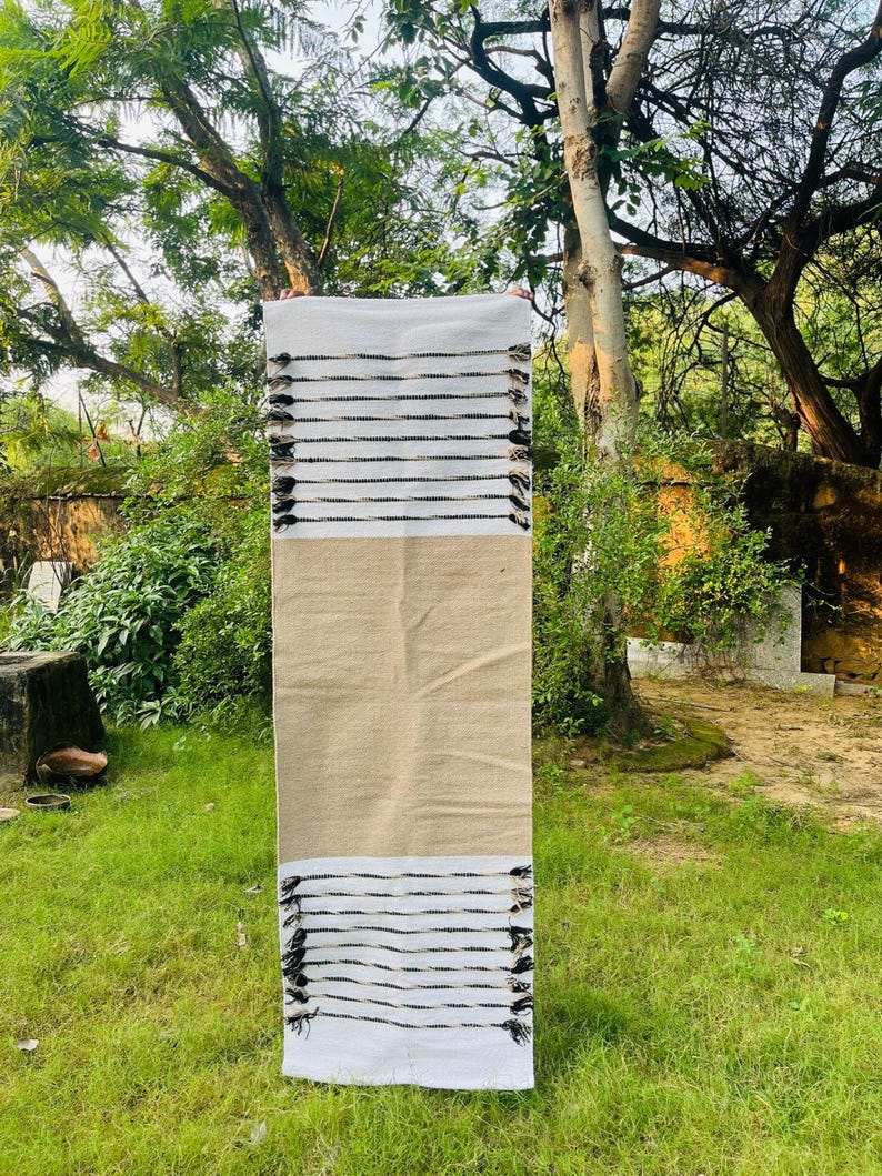 May include: A rectangular runner rug featuring a central solid tan section flanked by white ends. The white sections are decorated with horizontal black stripes and finished with small black tassels along the edges of the fabric.