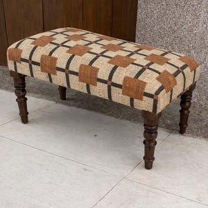 May include: A rectangular bench with a patterned seat and dark brown, turned legs. The seat has a repeating pattern of brown squares and black lines on a beige background. The legs are a dark, polished wood.