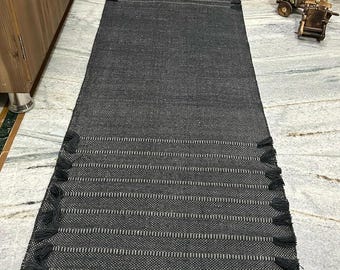 May include: A long, rectangular black runner rug featuring a woven texture with thin, horizontal white stripes spaced evenly throughout. The rug is finished with small, black decorative tassels attached along both long side edges.