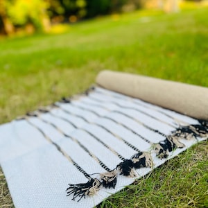 May include: A rectangular woven textile runner featuring a white base with thin, vertical black and beige stripes. The edges of the runner are finished with decorative, knotted fringe tassels in alternating black and beige thread colors.
