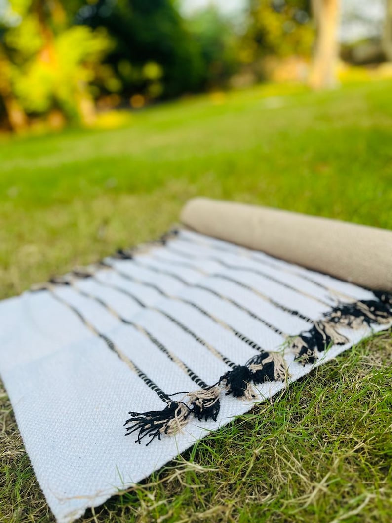 May include: A rectangular woven textile runner featuring a white base with thin, vertical black and beige stripes. The edges of the runner are finished with decorative, knotted fringe tassels in alternating black and beige thread colors.