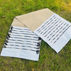 May include: A rectangular table runner featuring a central section of natural tan woven fabric flanked by two white end panels. The white sections are decorated with horizontal black stripes and finished with decorative black and tan fringe tassels.