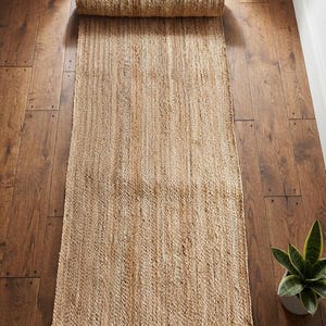 May include: A rectangular runner rug crafted from natural tan woven fibres, featuring a thick braided texture and a rolled end. The rug displays a consistent light brown colour with subtle variations in the natural material throughout its length.