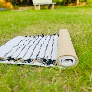 May include: A rectangular woven rug featuring a white base with thin, vertical black and tan stripes. The rug is partially rolled, revealing a textured tan underside, and is finished with decorative fringe along the edges of the flat section.