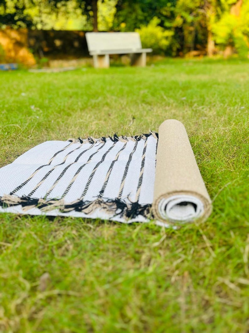 May include: A rectangular woven rug featuring a white base with thin, vertical black and tan stripes. The rug is partially rolled, revealing a textured tan underside, and is finished with decorative fringe along the edges of the flat section.