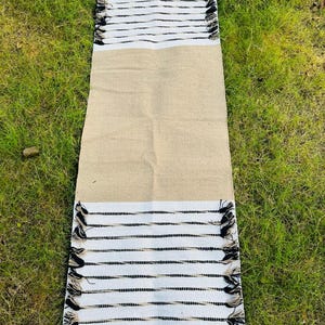 May include: A rectangular table runner featuring a solid tan center section flanked by white ends. Each white end displays horizontal black stripes and decorative fringe tassels along the edges, creating a textured and contrasting appearance.