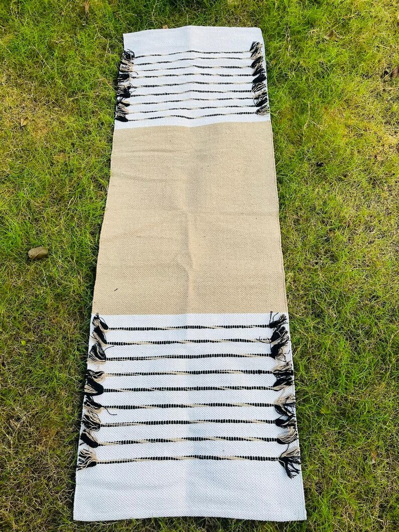 May include: A rectangular table runner featuring a solid tan center section flanked by white ends. Each white end displays horizontal black stripes and decorative fringe tassels along the edges, creating a textured and contrasting appearance.