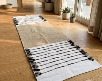 May include: A rectangular runner rug featuring a central tan jute section flanked by white ends with thin black horizontal stripes. The short edges are finished with decorative black and white knotted fringe tassels along the entire width.