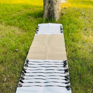 May include: A rectangular table runner featuring a central tan burlap section flanked by white ends. The white sections are decorated with horizontal black stripes and finished with small black tassels along the edges of the fabric.