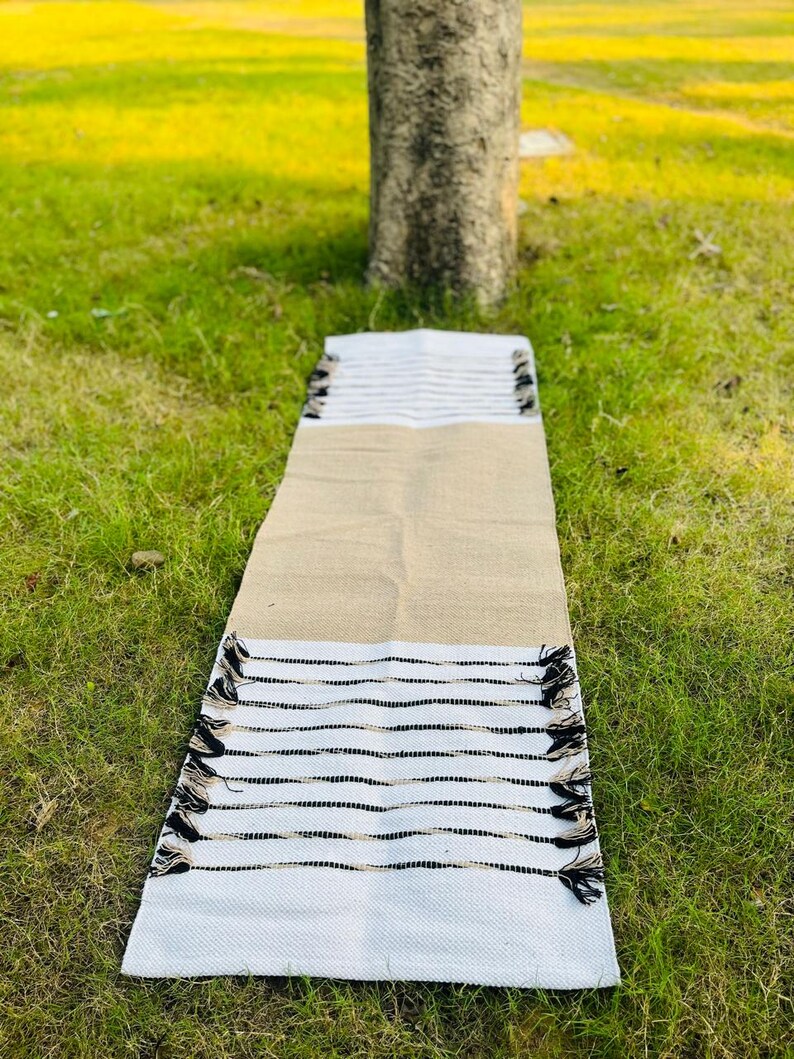 May include: A rectangular table runner featuring a central tan burlap section flanked by white ends. The white sections are decorated with horizontal black stripes and finished with small black tassels along the edges of the fabric.