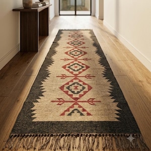May include: A long rectangular runner rug featuring a central beige field with a repeating geometric pattern in red and dark grey. The rug has a wide dark grey border and is finished with short, knotted fringe along the narrow ends.