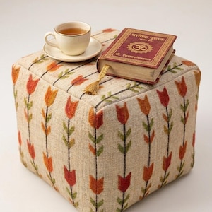 May include: A square ottoman with a floral pattern in orange, red, and green. A teacup and saucer sit on top, next to a book with gold lettering and a tassel bookmark. The ottoman is a neutral beige color.
