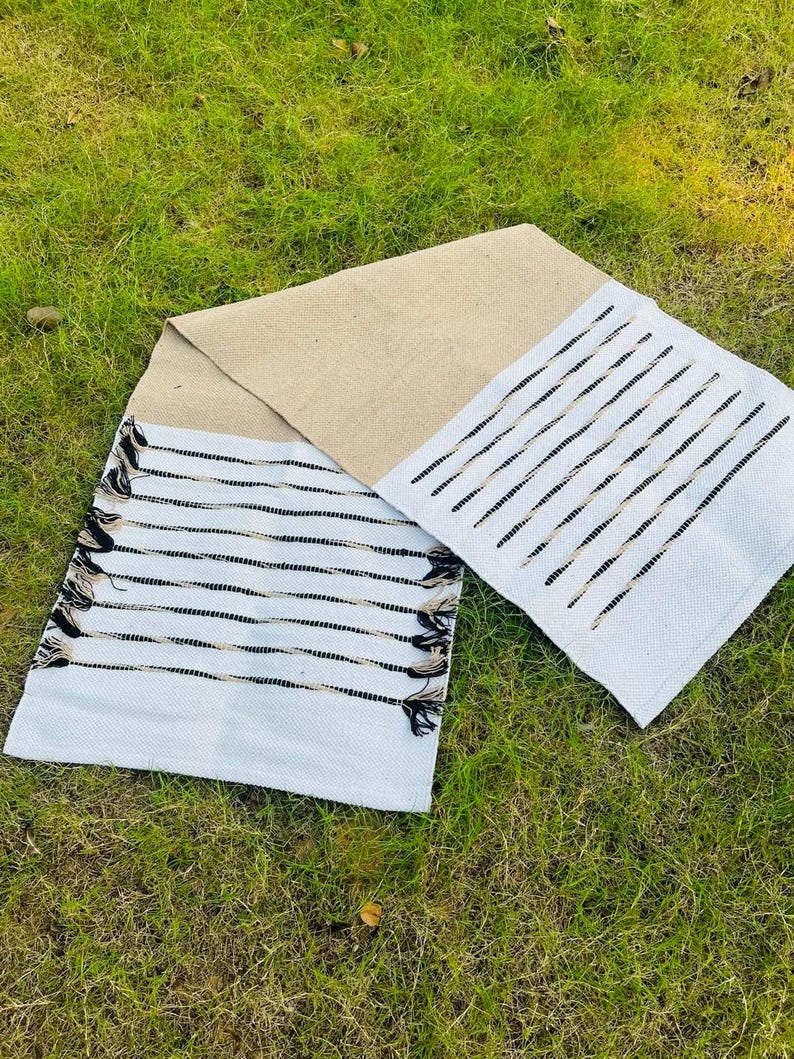 May include: A rectangular table runner featuring a central section of natural tan woven fabric flanked by two white end panels. The white sections are decorated with horizontal black stripes and finished with decorative black and tan fringe tassels.