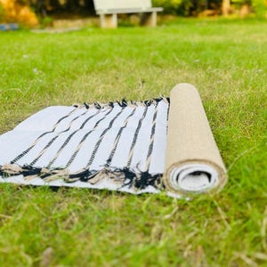 May include: A rectangular woven rug featuring a white base with thin, vertical black and tan stripes. The rug is partially rolled, revealing a textured tan underside, and is finished with decorative fringe along the edges of the flat section.
