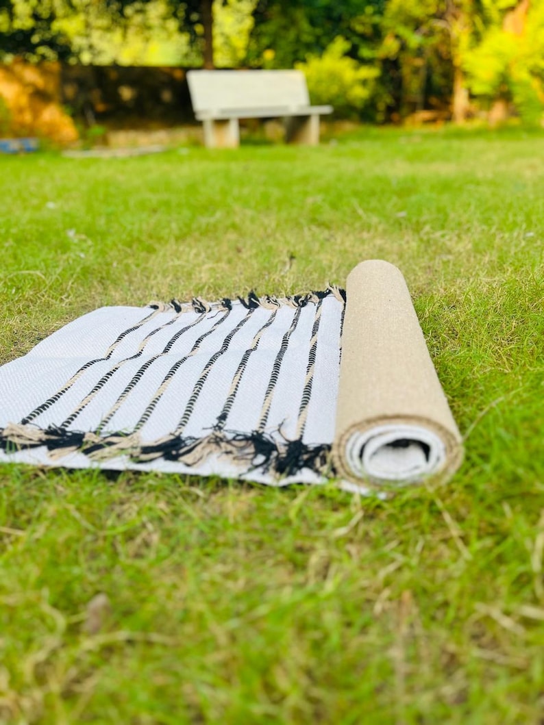 May include: A rectangular woven rug featuring a white base with thin, vertical black and tan stripes. The rug is partially rolled, revealing a textured tan underside, and is finished with decorative fringe along the edges of the flat section.