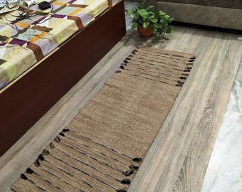 May include: A rectangular, woven jute rug with black stripes and fringe detailing. The rug is a natural tan color and is placed on a light wood-look floor. The rug is a home decor item.