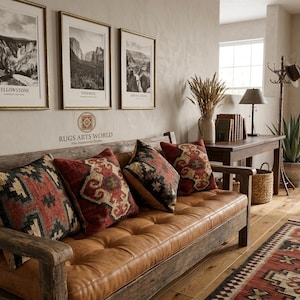 May include: A rustic living room scene featuring a wooden bench with a tufted, brown leather seat. Decorative pillows with red, black, and beige patterns rest on the bench. Three framed landscape prints hang above, and a patterned rug lies on the wooden floor.