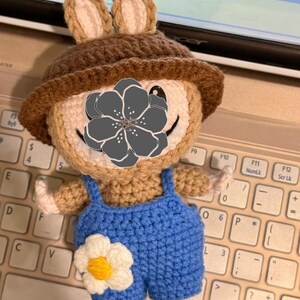 Peut inclure: Poup&eacute;e ours au crochet faite &agrave; la main, portant un chapeau marron et une salopette bleue. L'ours a une fleur blanche sur la salopette et est pos&eacute; sur un clavier.