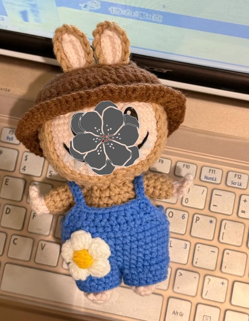Peut inclure: Poup&eacute;e ours au crochet faite &agrave; la main, portant un chapeau marron et une salopette bleue. L'ours a une fleur blanche sur la salopette et est pos&eacute; sur un clavier.
