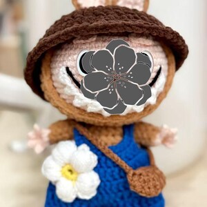 Peut inclure: Poup&eacute;e lapin au crochet faite &agrave; la main, portant un chapeau marron et une salopette bleue. La poup&eacute;e a une fleur blanche sur la salopette et un petit sac marron. Le lapin a un visage souriant.