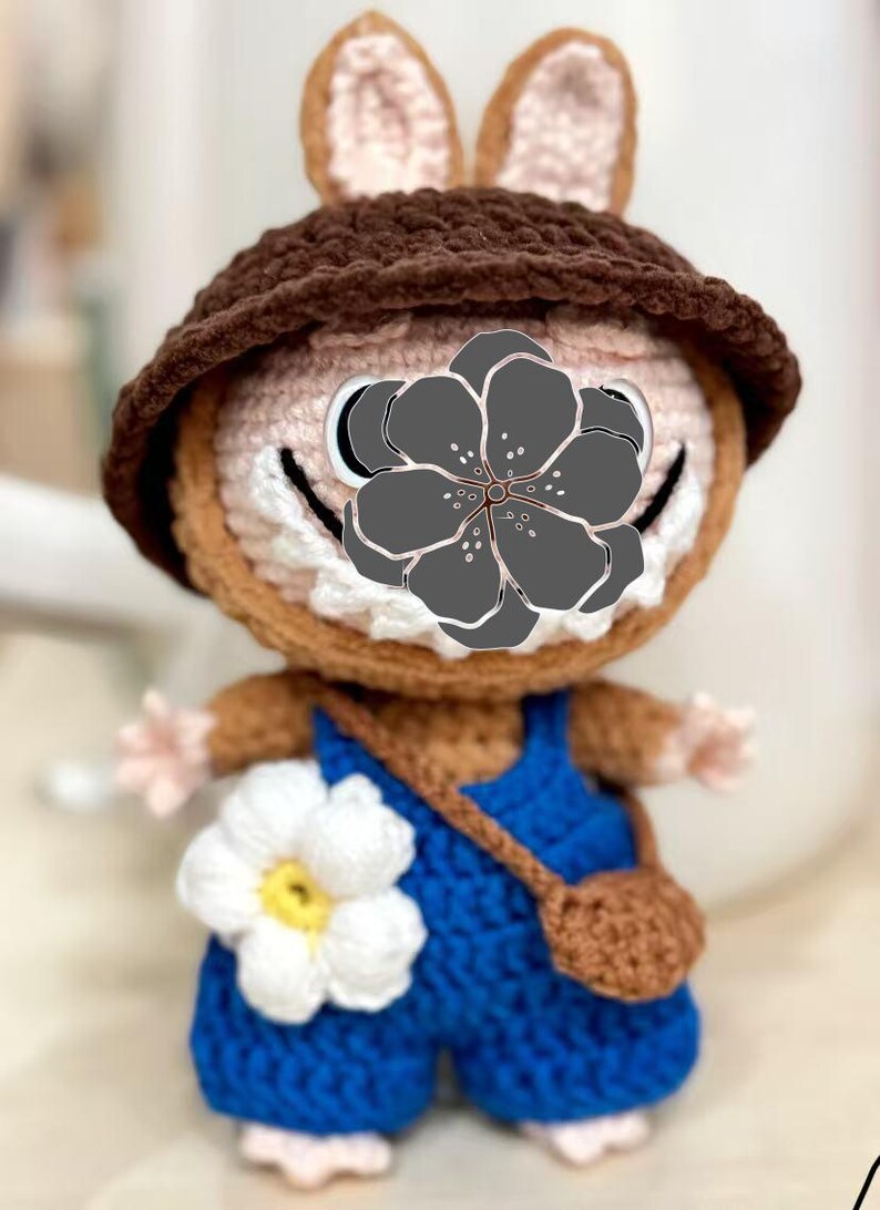 Peut inclure: Poup&eacute;e lapin au crochet faite &agrave; la main, portant un chapeau marron et une salopette bleue. La poup&eacute;e a une fleur blanche sur la salopette et un petit sac marron. Le lapin a un visage souriant.