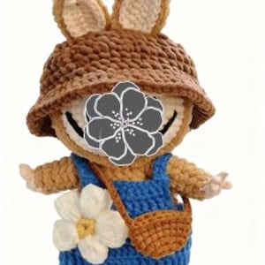 Peut inclure: Poup&eacute;e au crochet faite &agrave; la main, portant un chapeau cloche marron avec des oreilles de lapin, une salopette bleue et un petit sac marron. Une grande fleur blanche et jaune est attach&eacute;e &agrave; la salopette. La poup&eacute;e a un visage souriant.