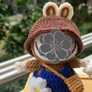 Peut inclure: Poup&eacute;e grenouille au crochet faite &agrave; la main, portant un chapeau marron avec des oreilles cr&egrave;me, une salopette bleue et une petite sacoche marron. La poup&eacute;e a des bras blancs et une fleur blanche sur sa salopette. Elle est expos&eacute;e sur un socle en bois.