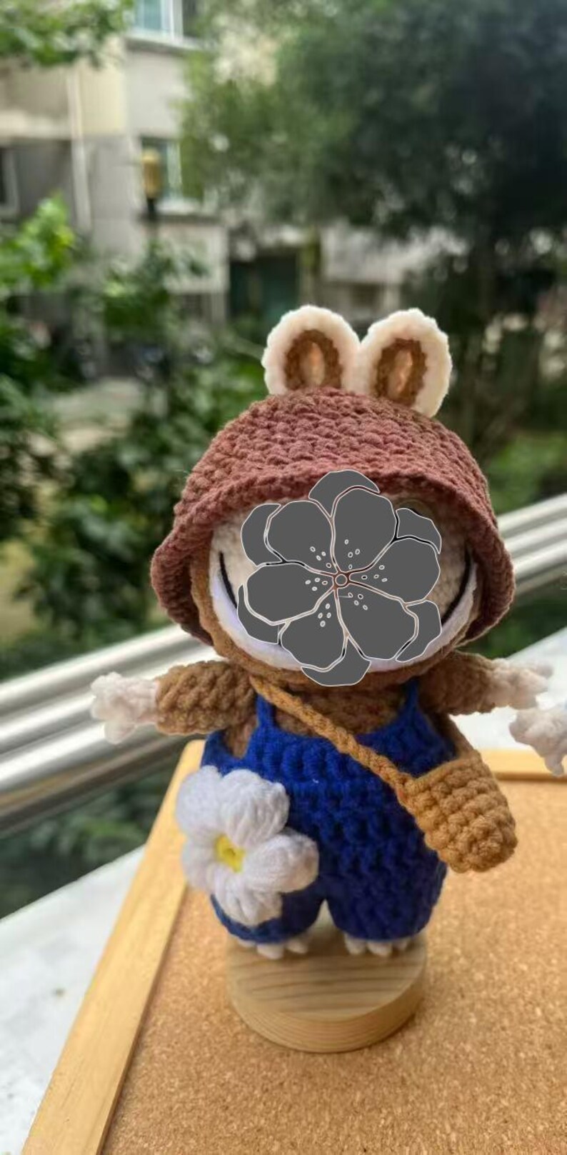 Peut inclure: Poup&eacute;e grenouille au crochet faite &agrave; la main, portant un chapeau marron avec des oreilles cr&egrave;me, une salopette bleue et une petite sacoche marron. La poup&eacute;e a des bras blancs et une fleur blanche sur sa salopette. Elle est expos&eacute;e sur un socle en bois.