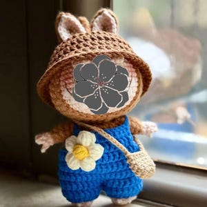 Peut inclure: Poup&eacute;e lapin au crochet faite &agrave; la main, portant un chapeau marron et une salopette bleue. La poup&eacute;e a une fleur blanche et jaune sur la salopette et un petit sac beige. Le lapin a une apparence mignonne et amicale.